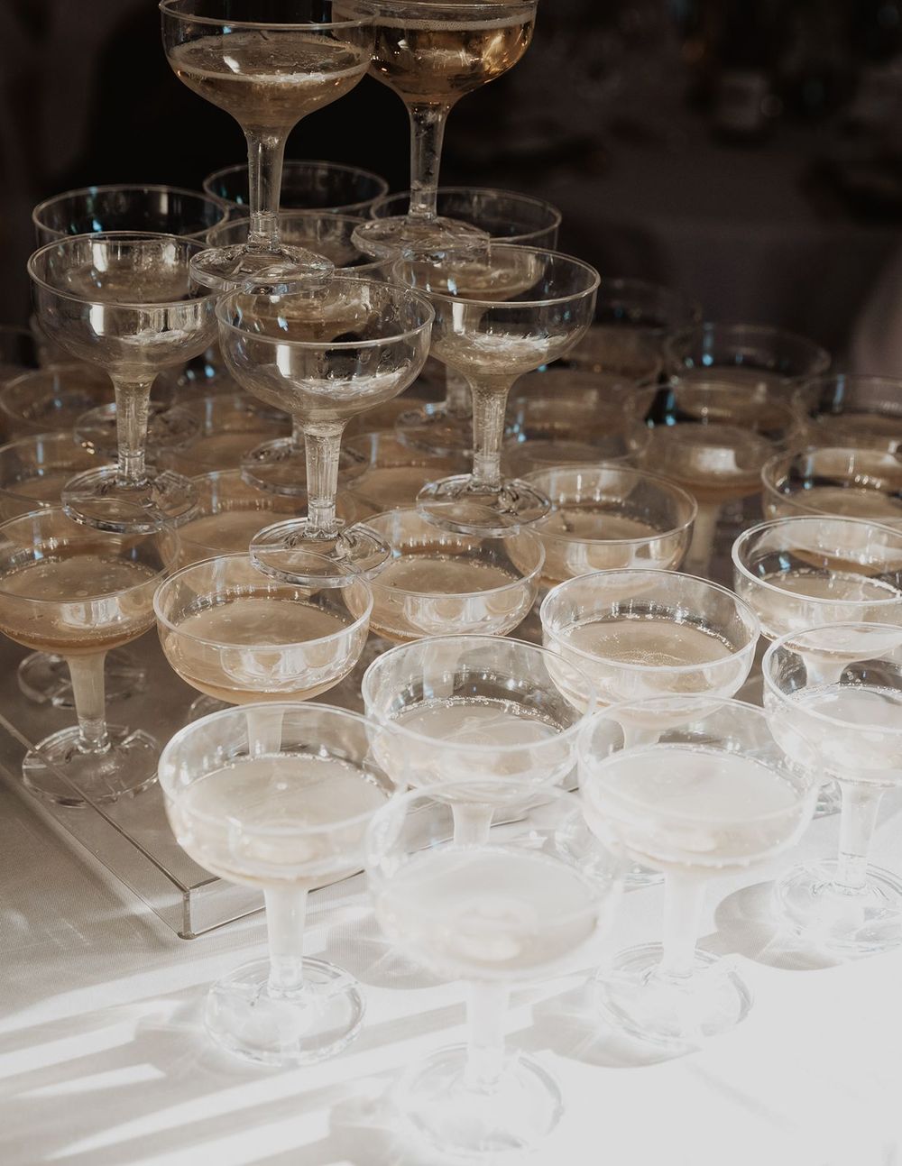 Pyramid of champagne coupe glasses arranged on a white tablecloth at a wedding reception.
