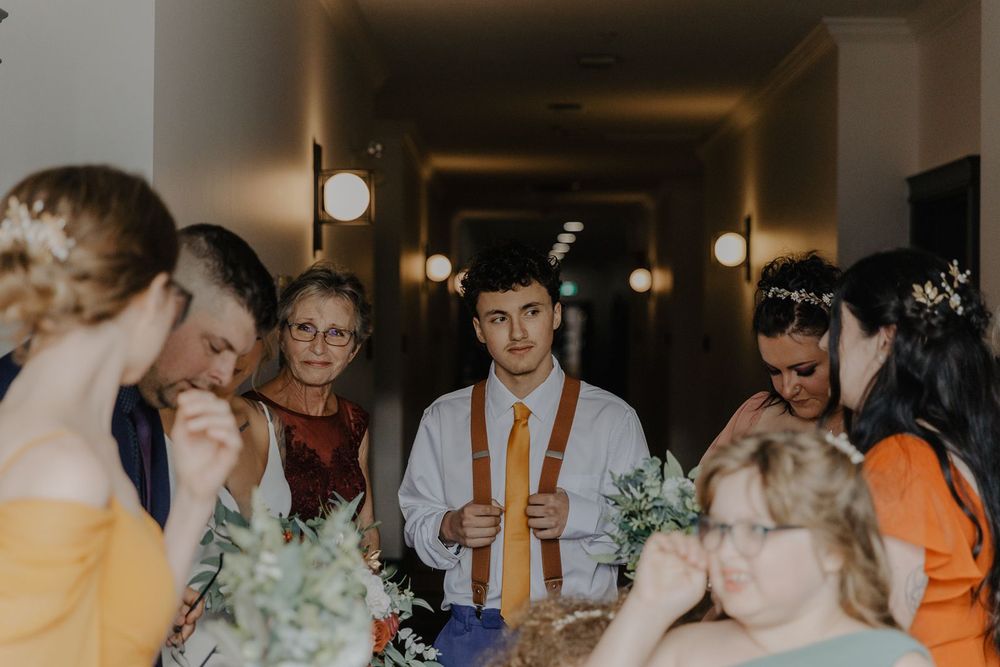 A groom in suspenders stands in a hallway surrounded by bridesmaids holding floral bouquets.