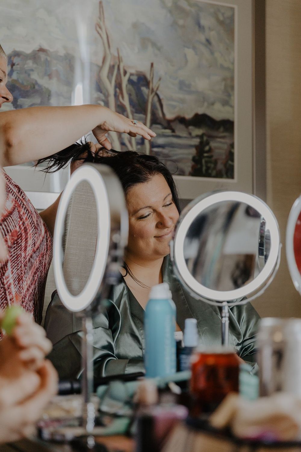 A bride smiles as a stylist styles her dark hair using a lighted vanity mirror.