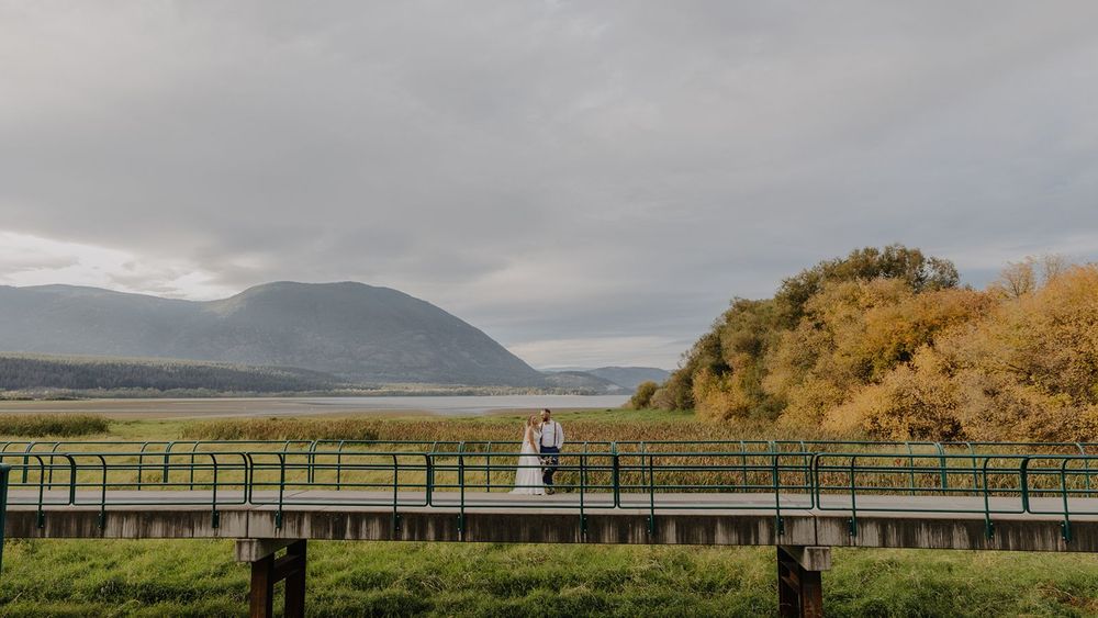 A bride and groom stand together on a wooden bridge with a scenic mountain lake backdrop.