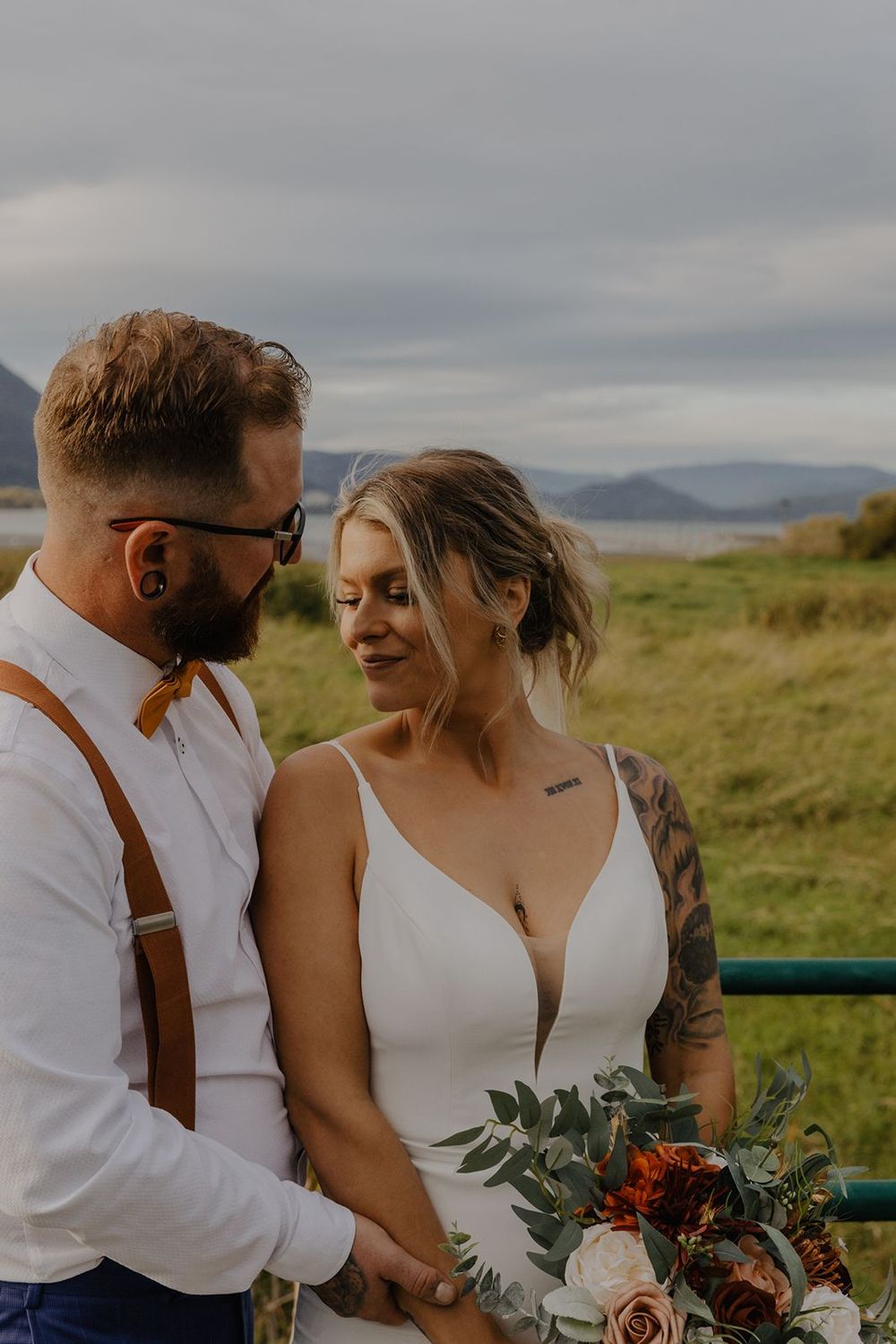 Bride with tattoos holding bouquet smiles at tattooed groom in suspenders outdoors near water.