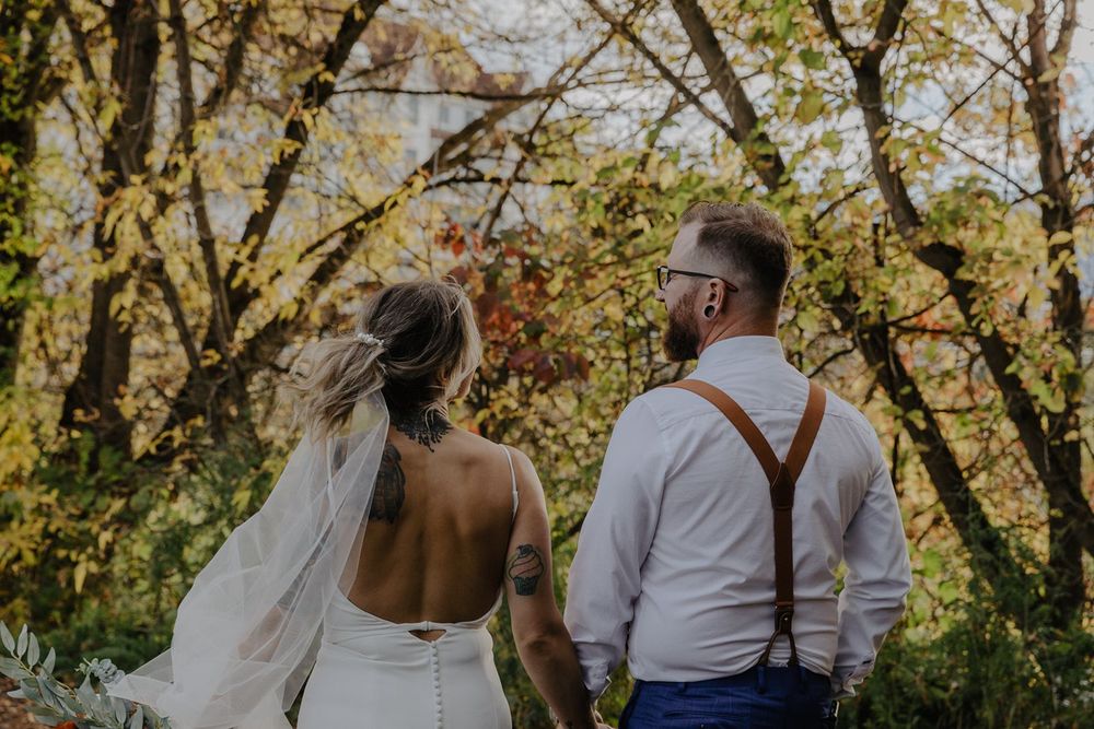 Wedding couple walks hand in hand through autumn forest, bride showing back tattoo in open gown.