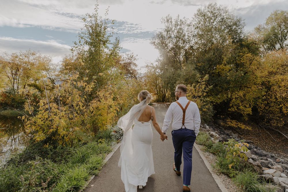 Bride and groom walk hand in hand down path surrounded by golden autumn foliage near river.