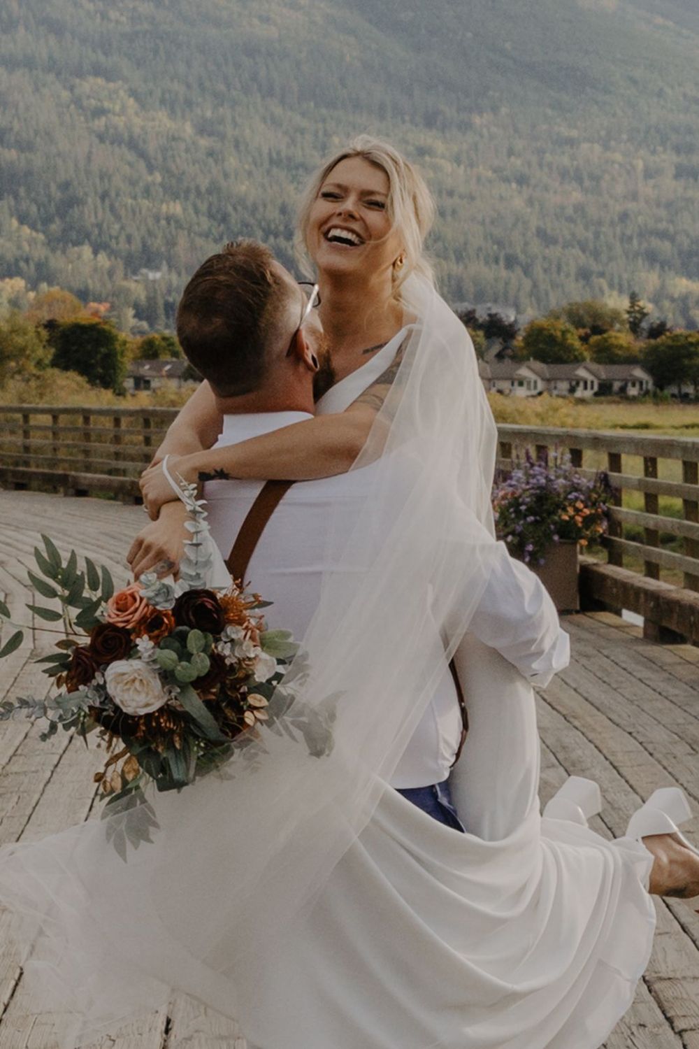 Laughing bride lifted by groom on wooden deck with mountain backdrop and bridal bouquet.