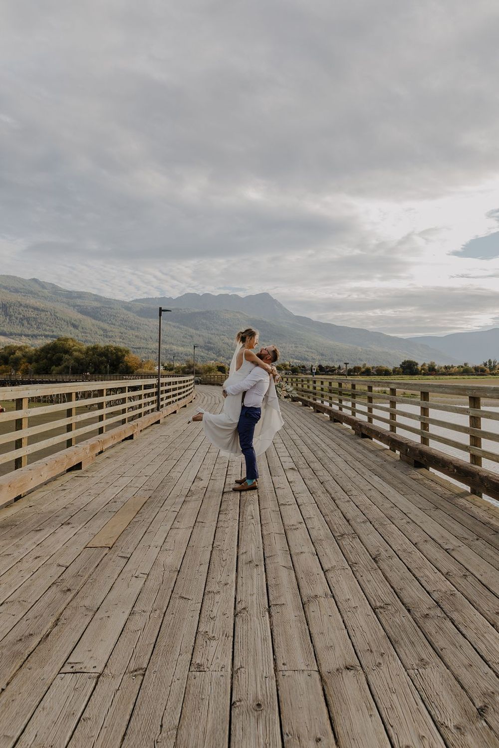 Groom lifts bride on rustic wooden pier with dramatic mountain and cloudy sky backdrop.