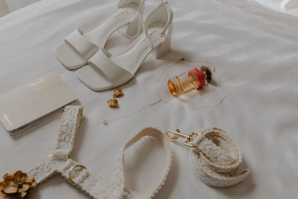 Flat lay of white bridal accessories including heeled sandals, lace garter, perfume, and gold jewelry on white fabric.