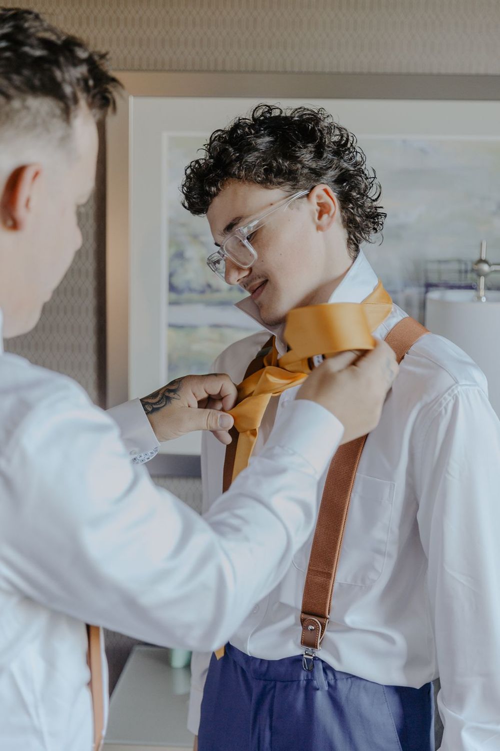 Person helping groom tie golden yellow necktie with brown suspenders during wedding day preparations.