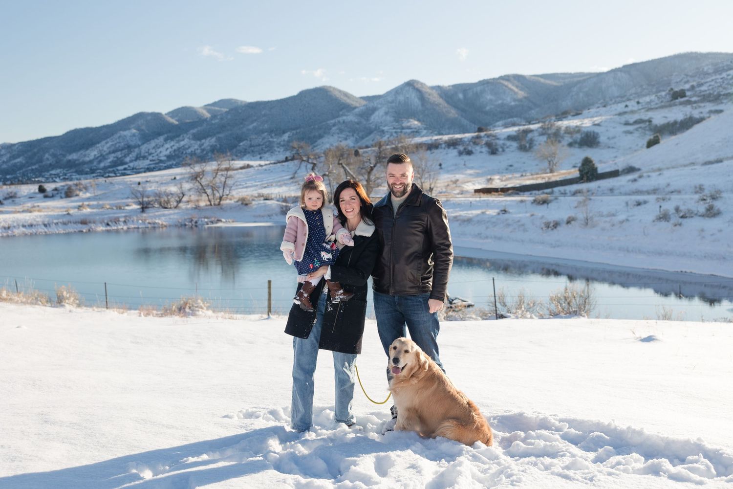 Three people and a dog pose together on a snowy hillside overlooking a scenic mountain lake in winter.