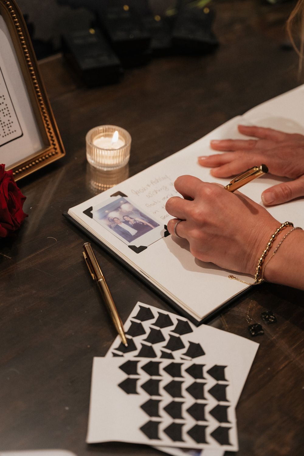 A person writes in a guestbook with a candle and decorative black heart designs on a dark wooden table.