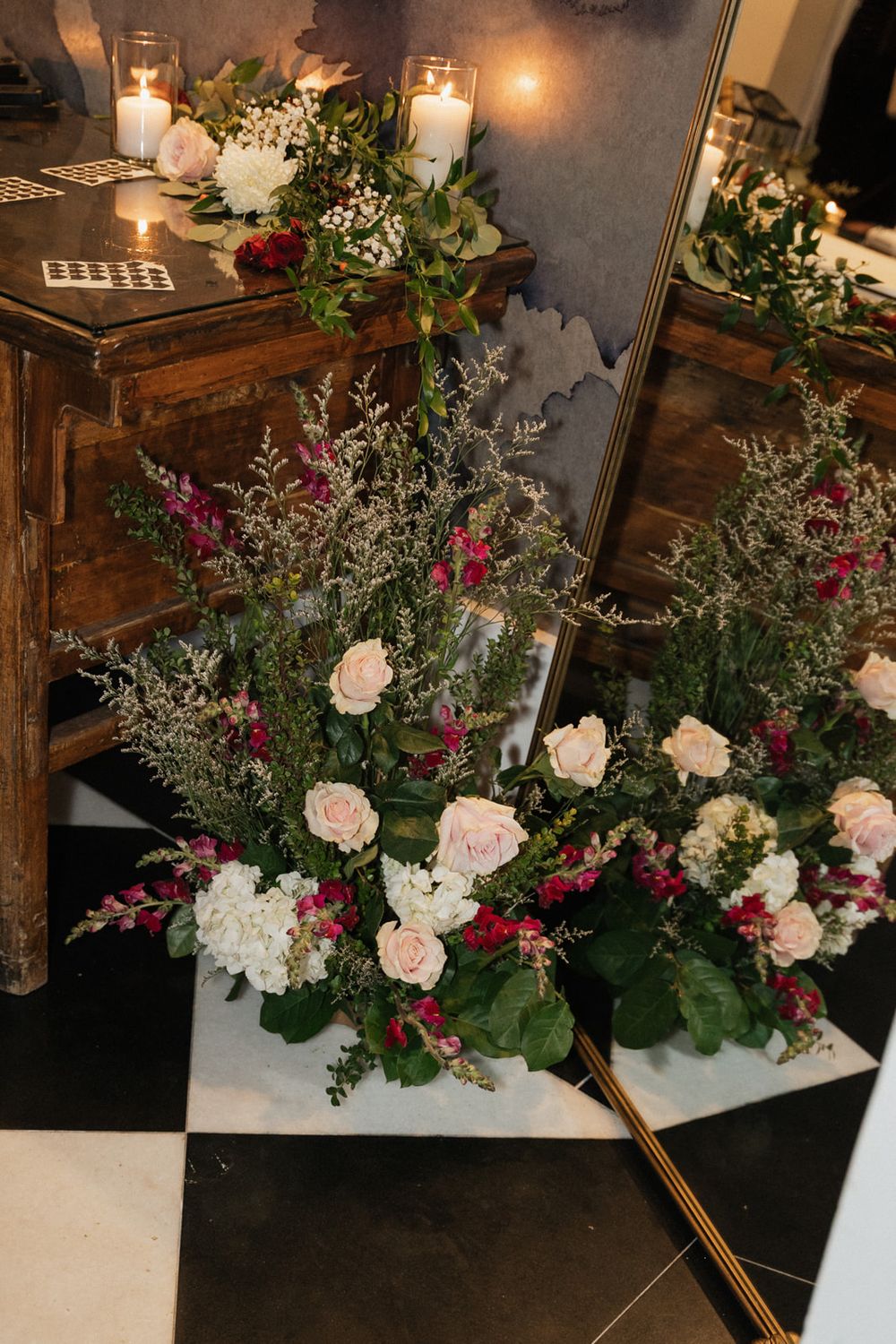 Elegant floral centerpiece with pink roses, white hydrangeas and greenery adorned with glowing candles on a checkered floor.