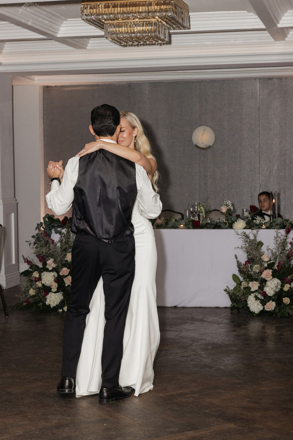 A couple in formal wedding attire dance together in an elegant ballroom with floral decorations and chandeliers.