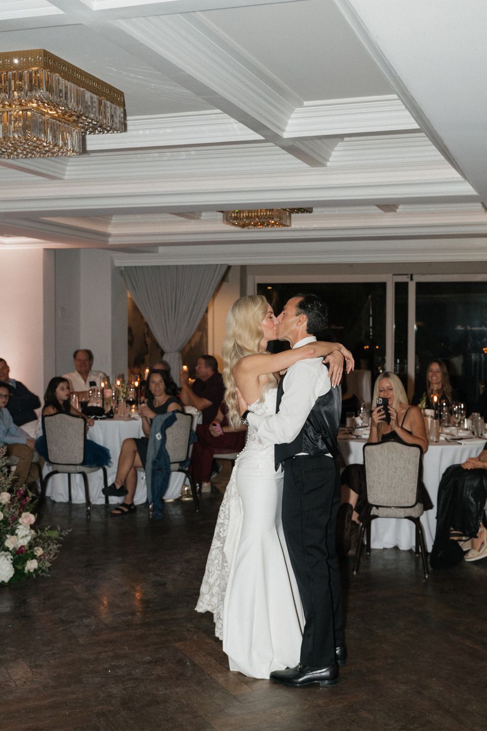 Wedding reception guests dancing on a dark wooden dance floor under elegant crystal chandeliers.