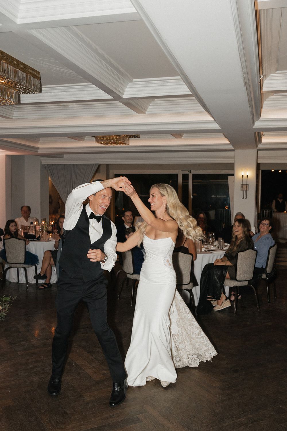Wedding guests twirl and dance together on a dark wooden dance floor at an elegant indoor reception venue.
