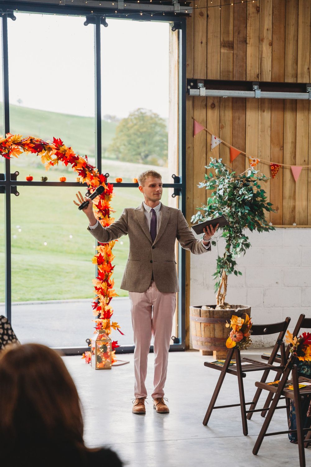 Wedding ceremony setup in a rustic barn venue with autumn-themed orange floral arch and wooden chairs.