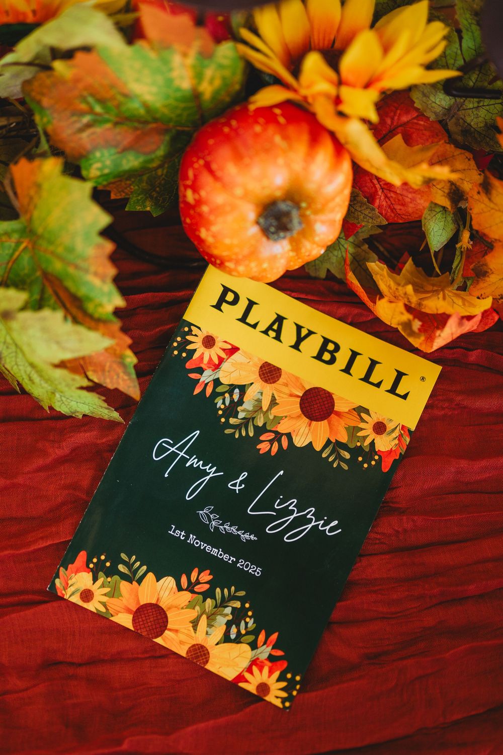 Fall-themed wedding program with playbill design surrounded by orange and yellow flowers on burgundy fabric.