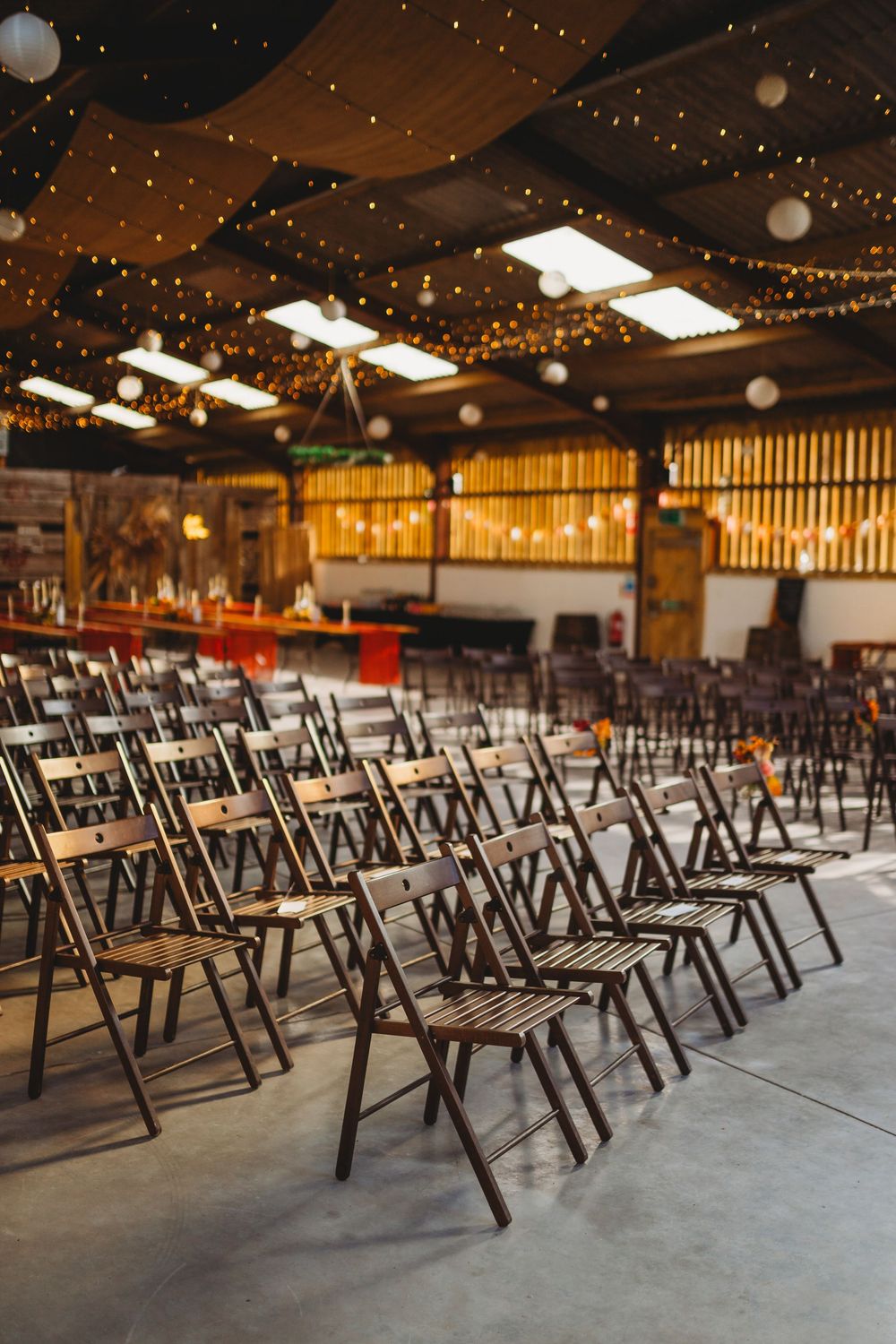 Rows of wooden folding chairs arranged in a rustic barn venue with twinkling string lights overhead.