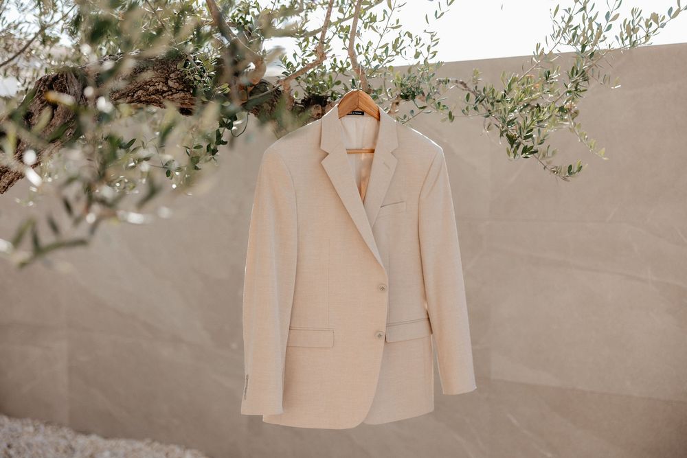 A light beige linen blazer jacket hangs on a wooden hanger against a textured gray wall with green foliage accents.