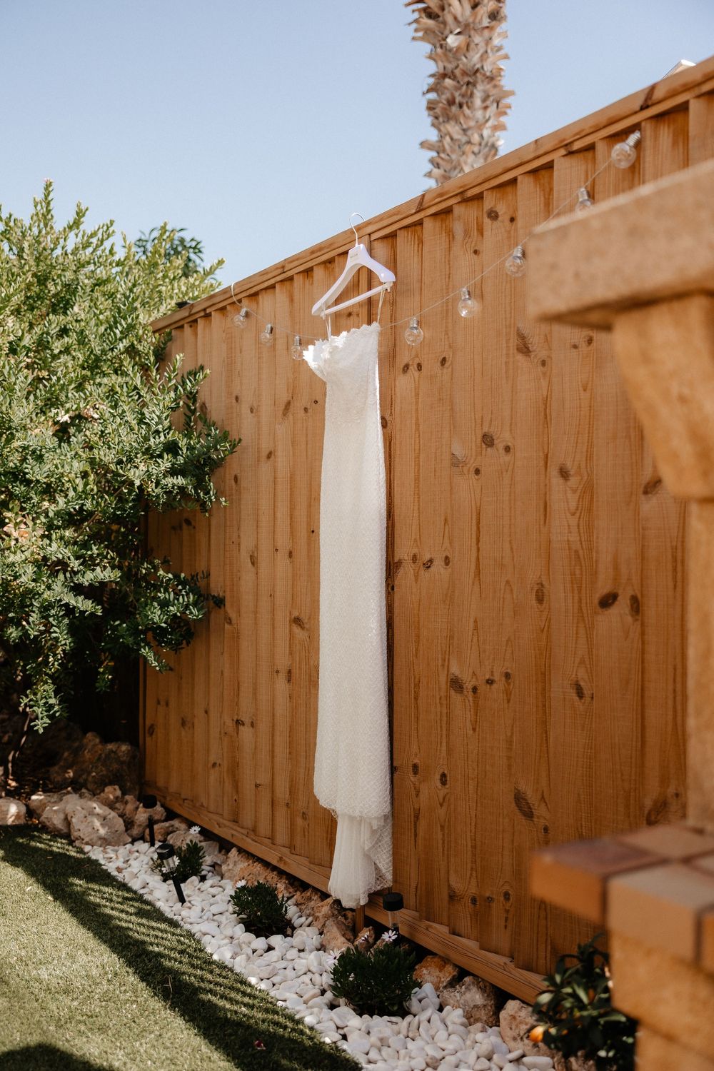 A white wedding dress hangs elegantly against a wooden fence with decorative rocks and greenery in the background.