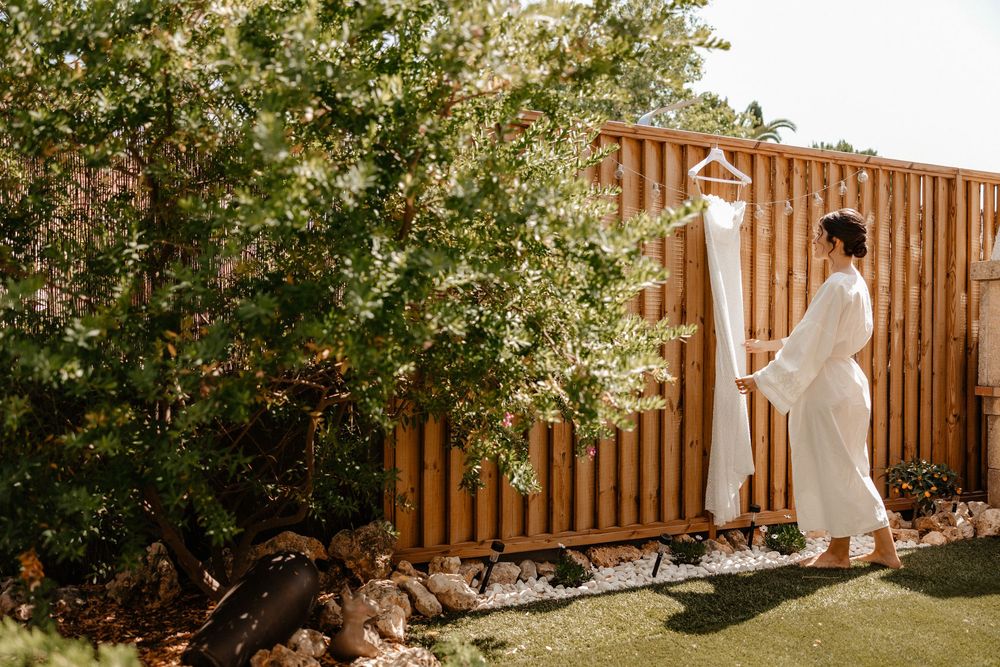 A person in a flowing white dress stands by a wooden fence in a garden with blooming spring flowers.