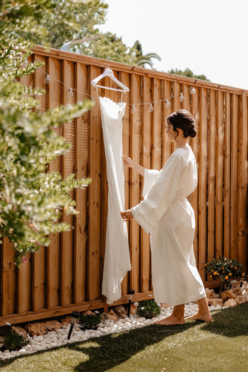 A white wedding dress hanging on a wooden fence with sunlit greenery in the background, shown in a series of photos.