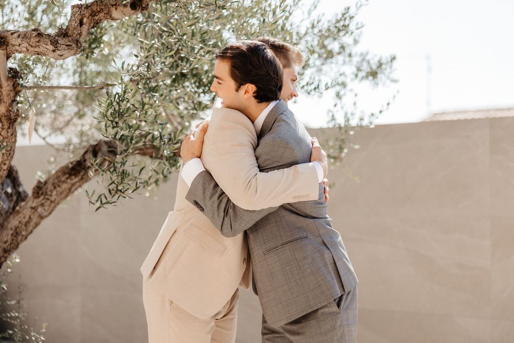 Couple sharing an intimate embrace in front of a beige wall with olive trees in the background.