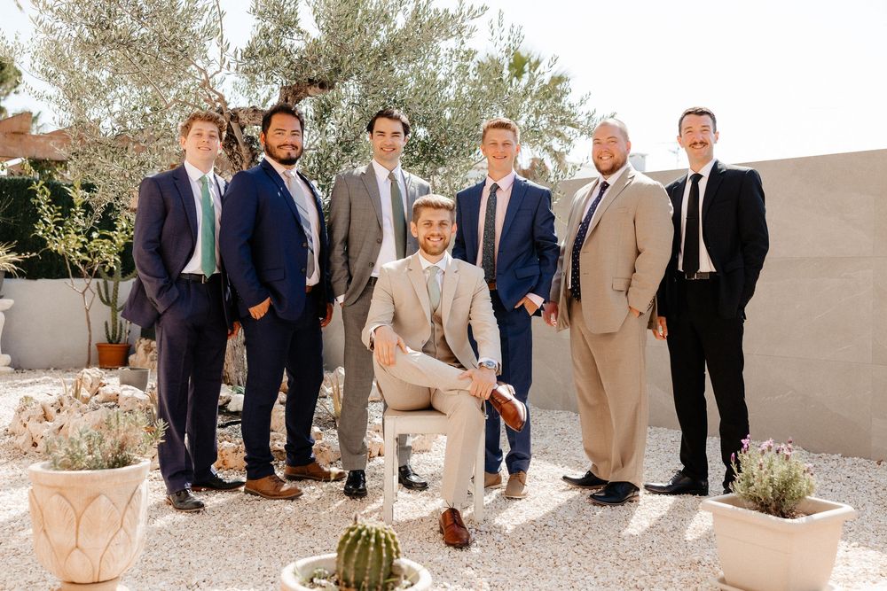 Group of people in formal attire standing outdoors against a light colored wall with desert plants and landscaping.