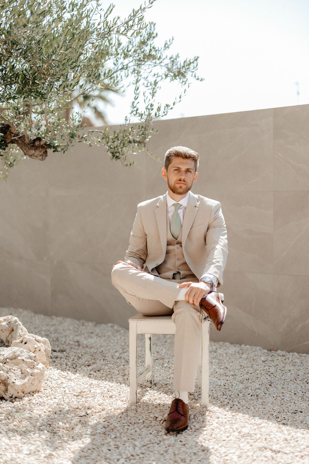 A person in a light beige suit sits elegantly on a white chair against a neutral wall with greenery peeking overhead.
