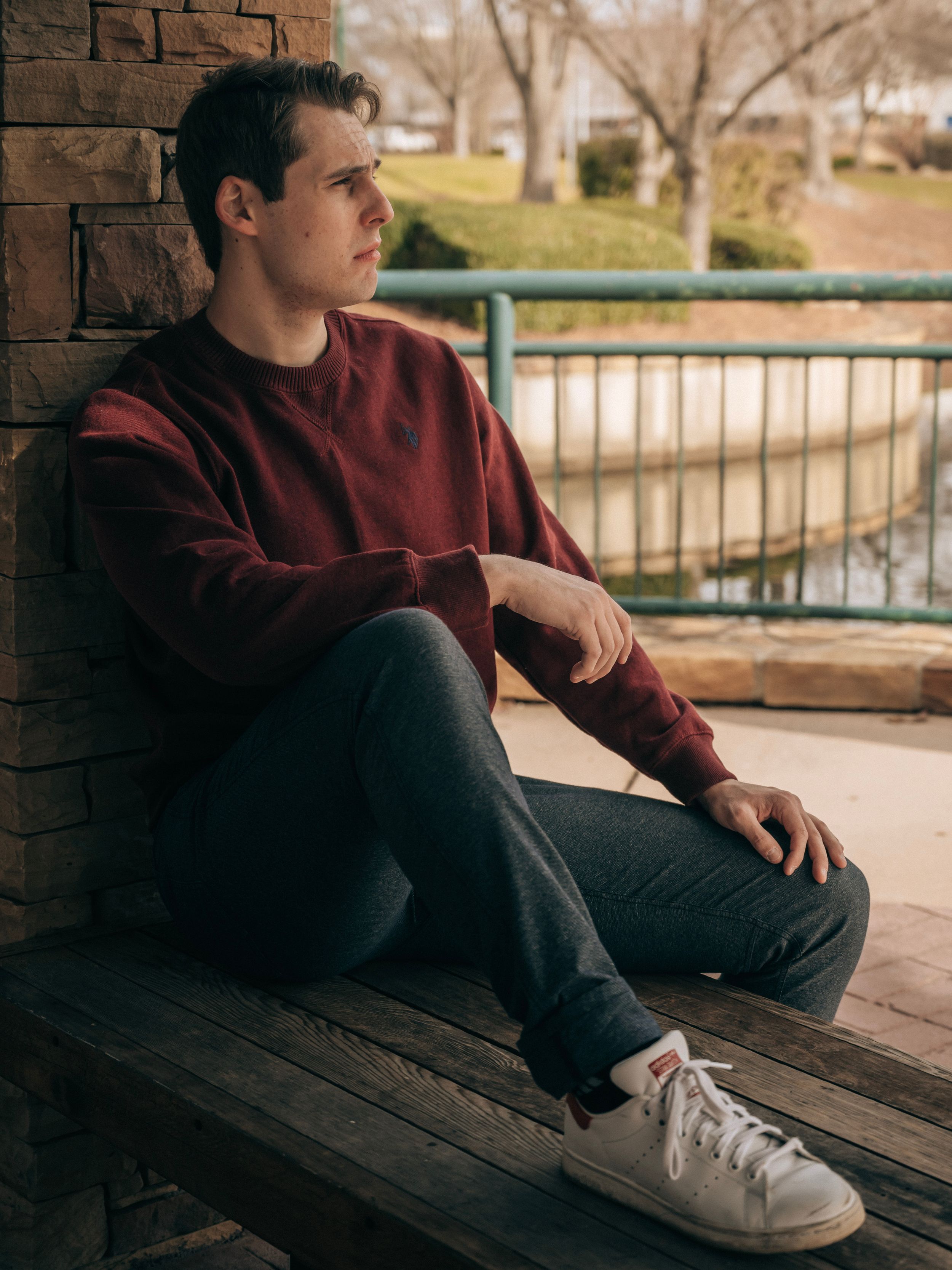Person in burgundy sweater sits contemplatively on wooden bench near brick wall and railing.