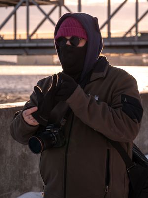 Photographer in winter gear and beanie takes photos during sunset.