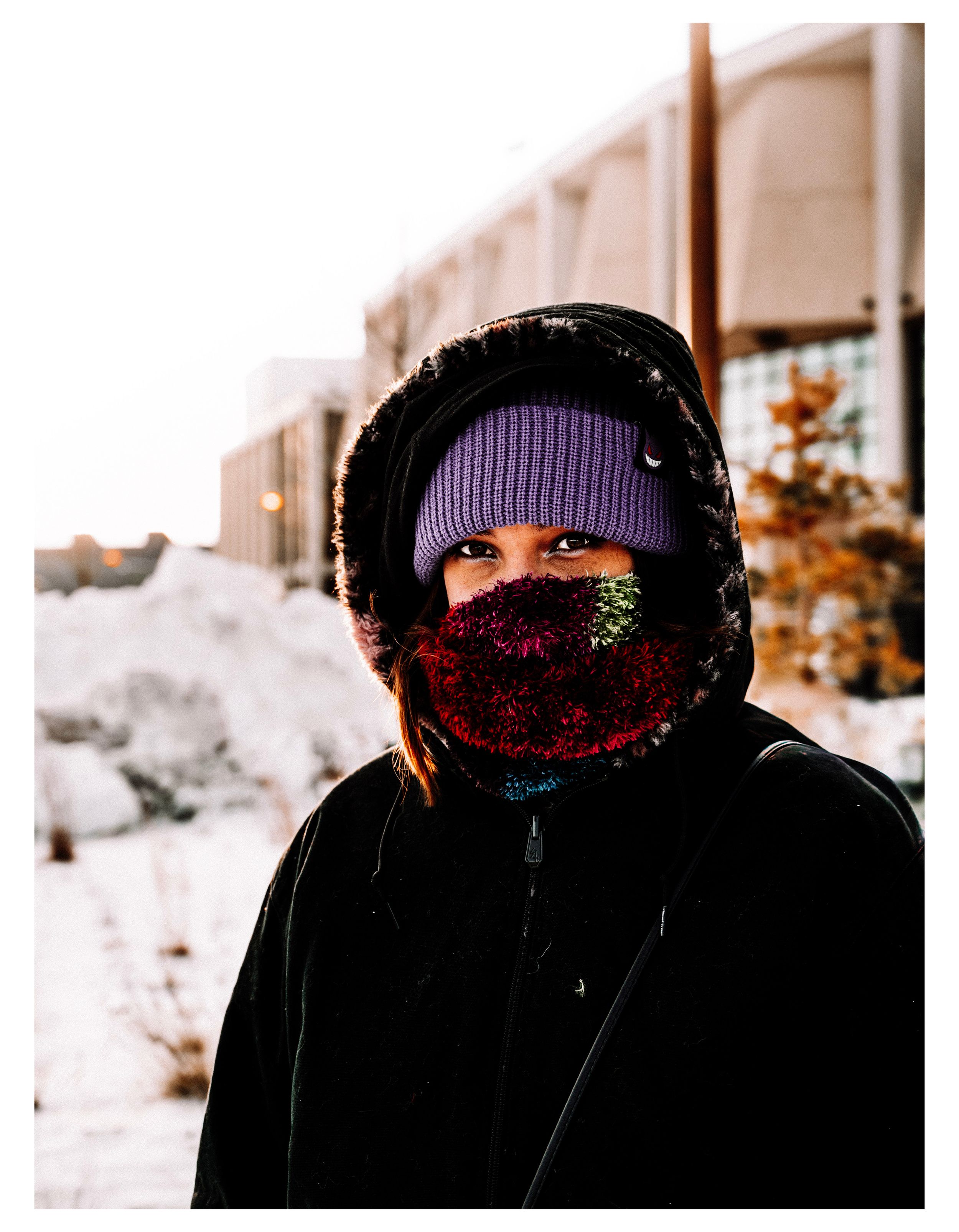 Person bundled in winter clothing and purple beanie stands against snowy background.