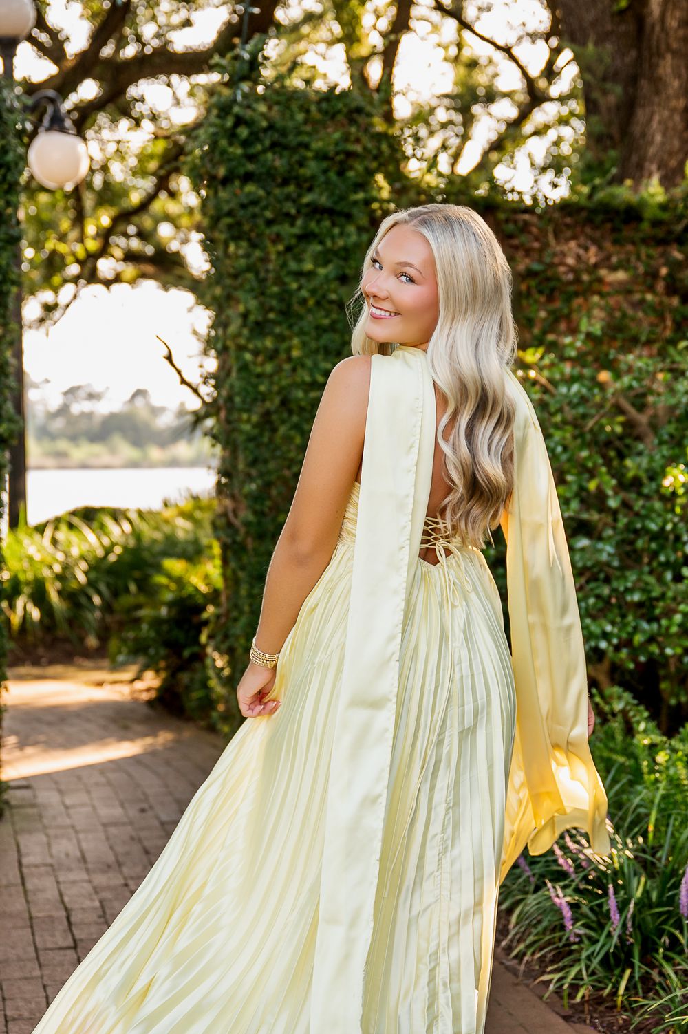 Light yellow pleated maxi dress showcased on brick pathway with garden backdrop.