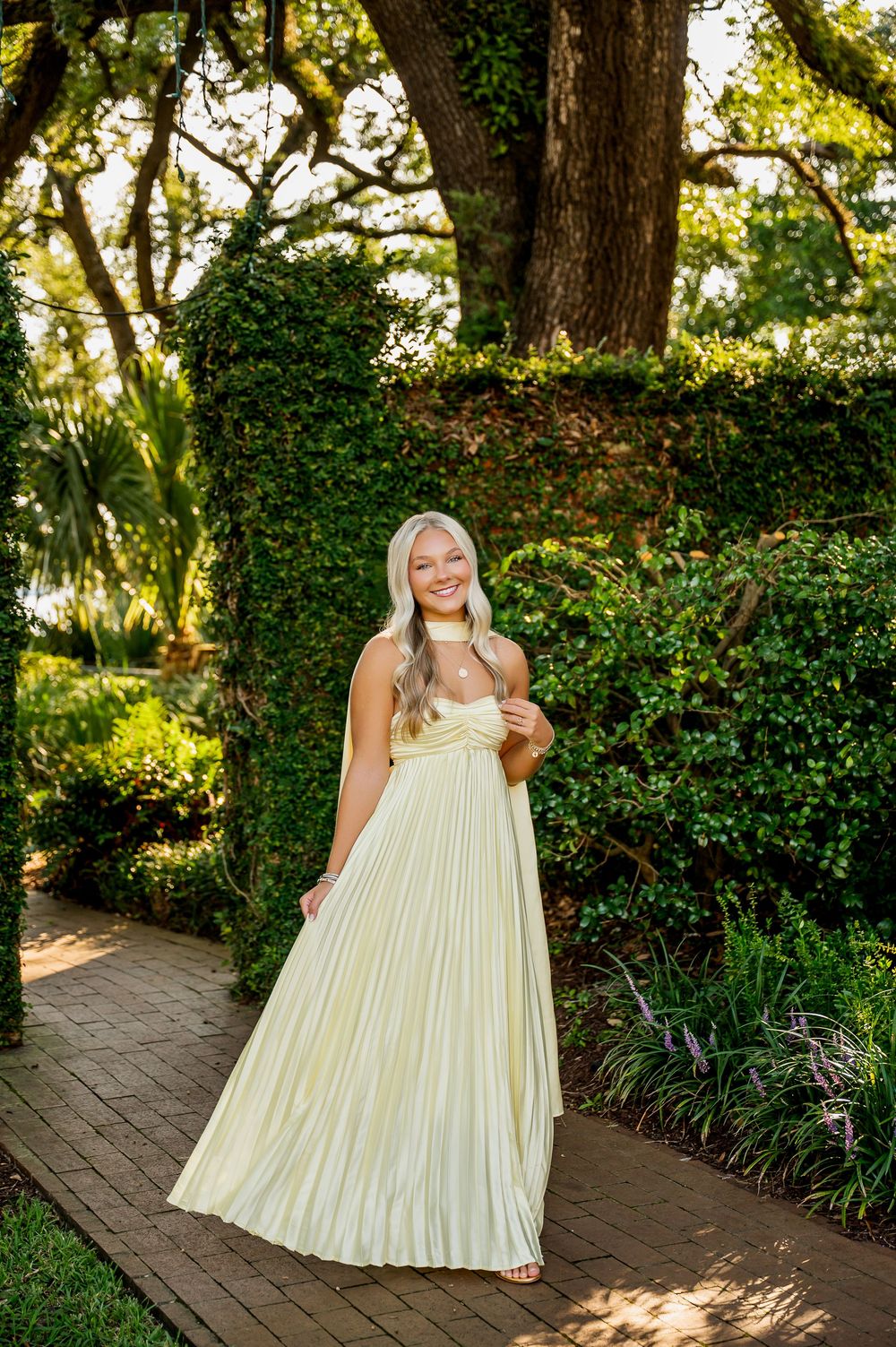 Flowing pale yellow pleated dress captured on brick pathway surrounded by green garden foliage.