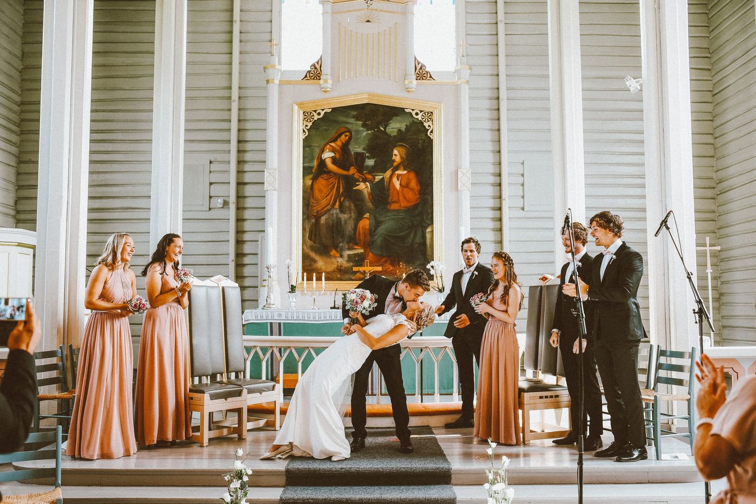 A wedding ceremony taking place in a church with bridesmaids in peach dresses and an ornate altar painting.
