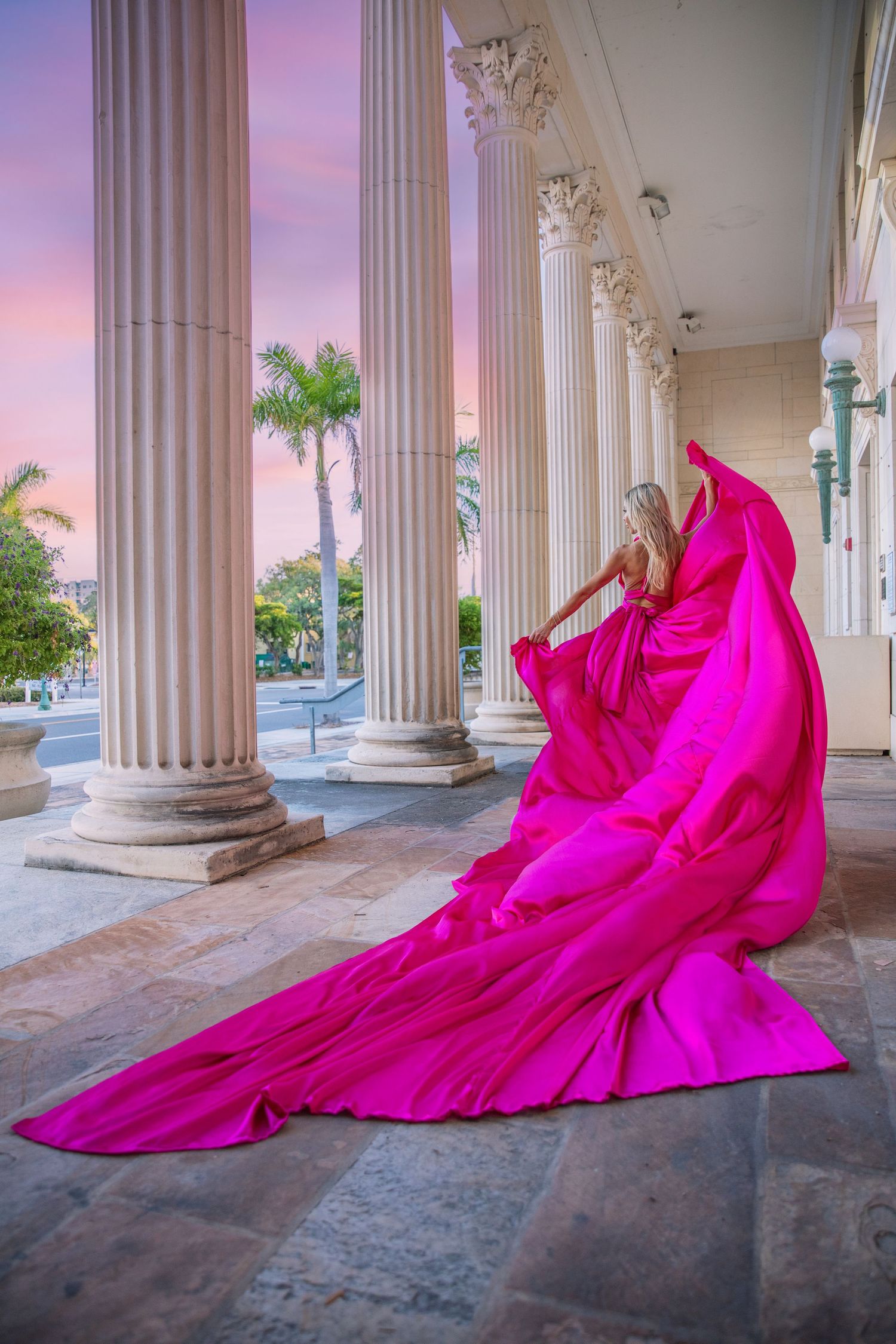 A vibrant hot pink flowing dress fabric cascades dramatically between classical white columns at sunset.
