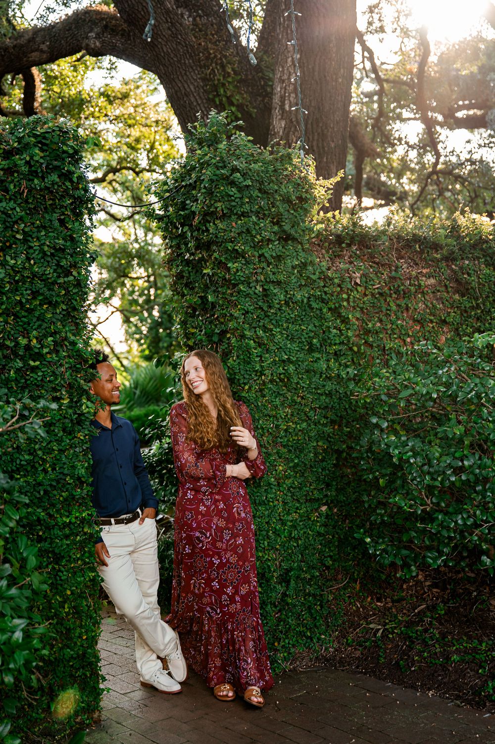 A couple shares a playful moment in a garden setting with lush greenery and trees.