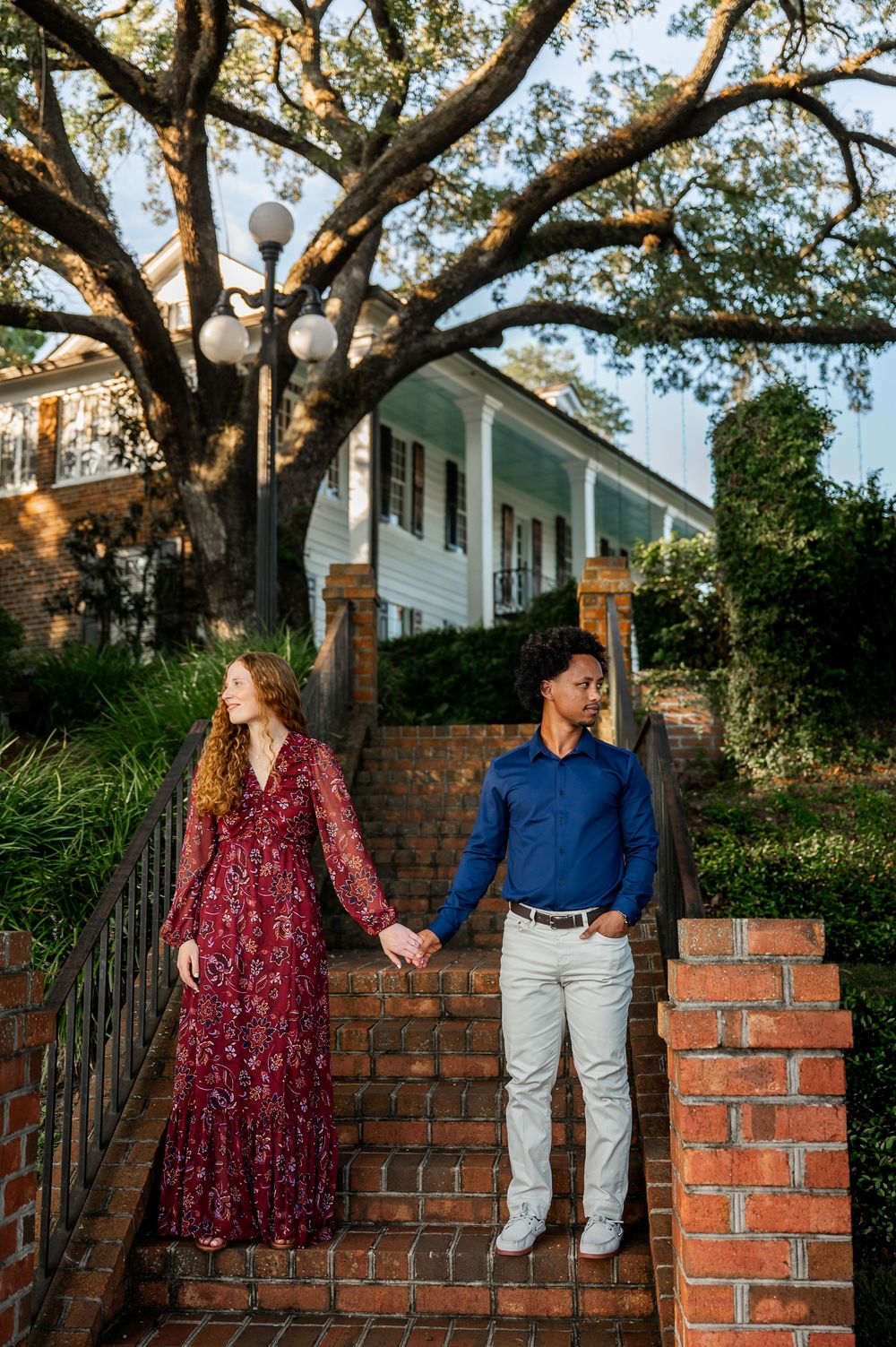 A couple poses along a brick wall with white houses and trees in the background.