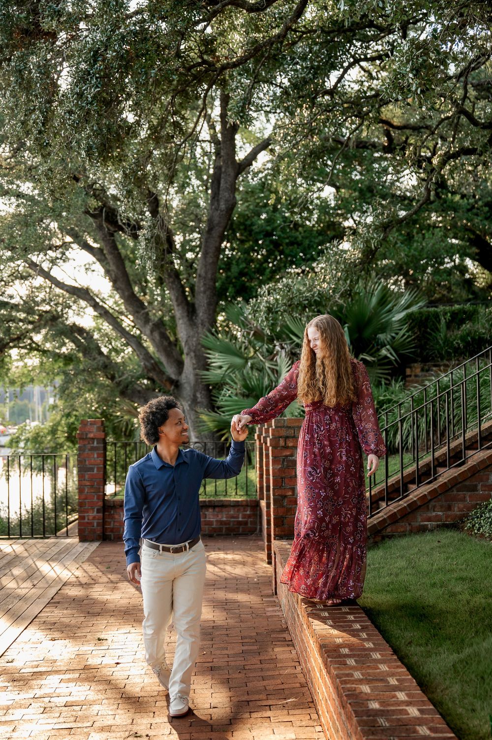 A couple walks and poses along a brick-lined garden path under large oak trees.