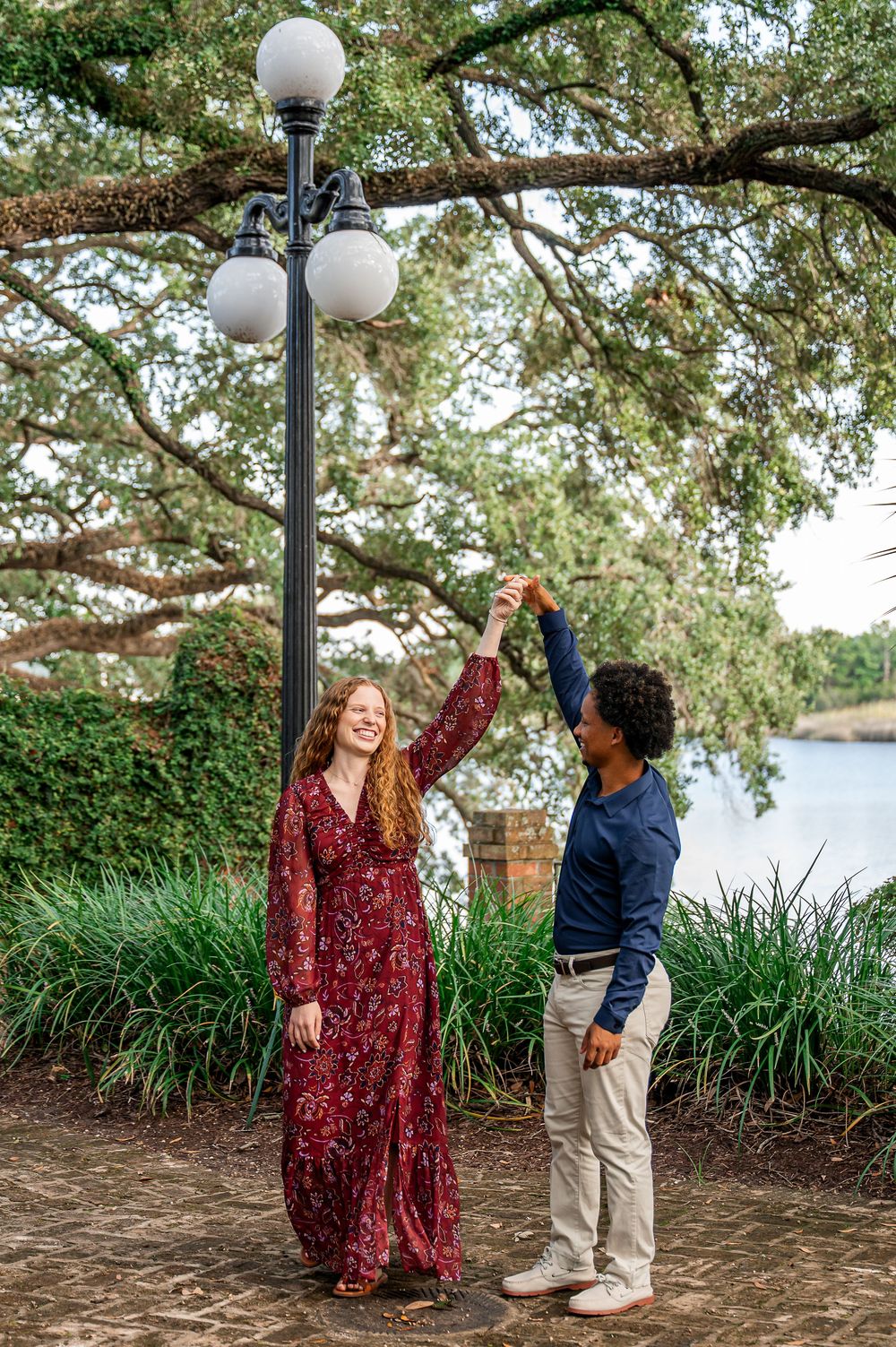 A couple shares a playful moment under trees with white globe lamps in a park setting.