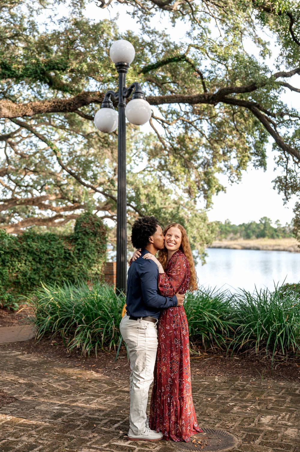 A romantic couple poses together near ornamental grasses and white lamp posts by the water.