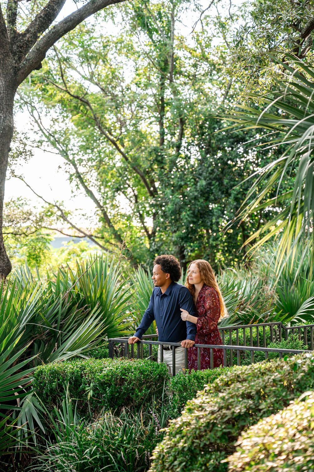 A couple walks together through a lush garden path with palm fronds and greenery.