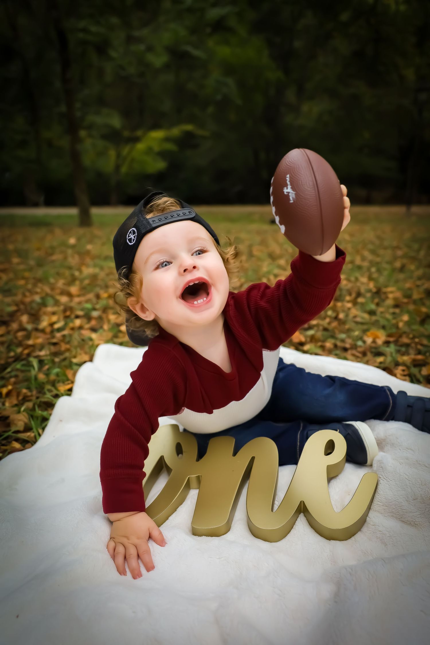 Baby in burgundy sweater playing with gold letters on white fabric outdoors among fallen leaves.