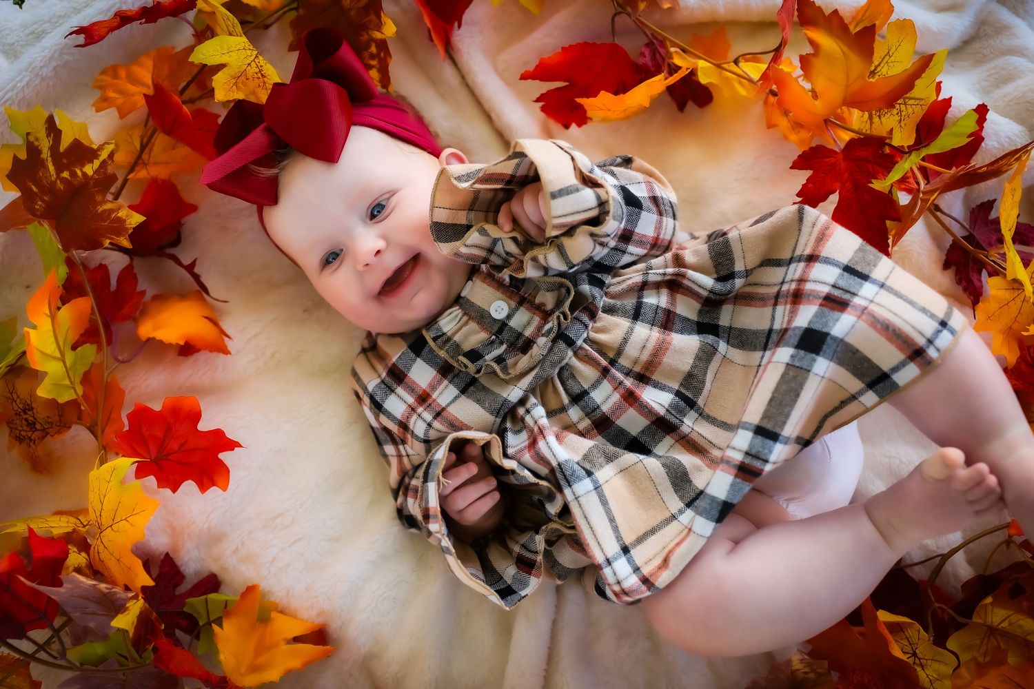 Baby lying among colorful autumn leaves wearing plaid clothing.
