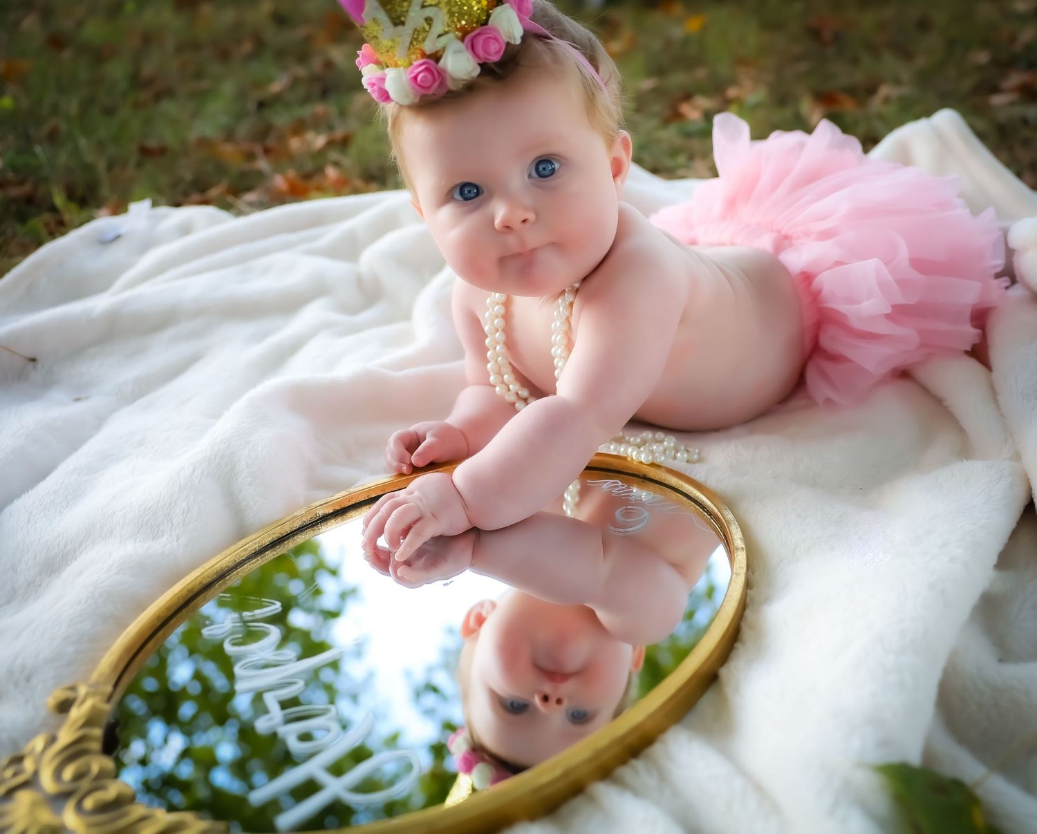 Baby in pink tulle lying on white fabric with floral decorations and a gold mirror frame.