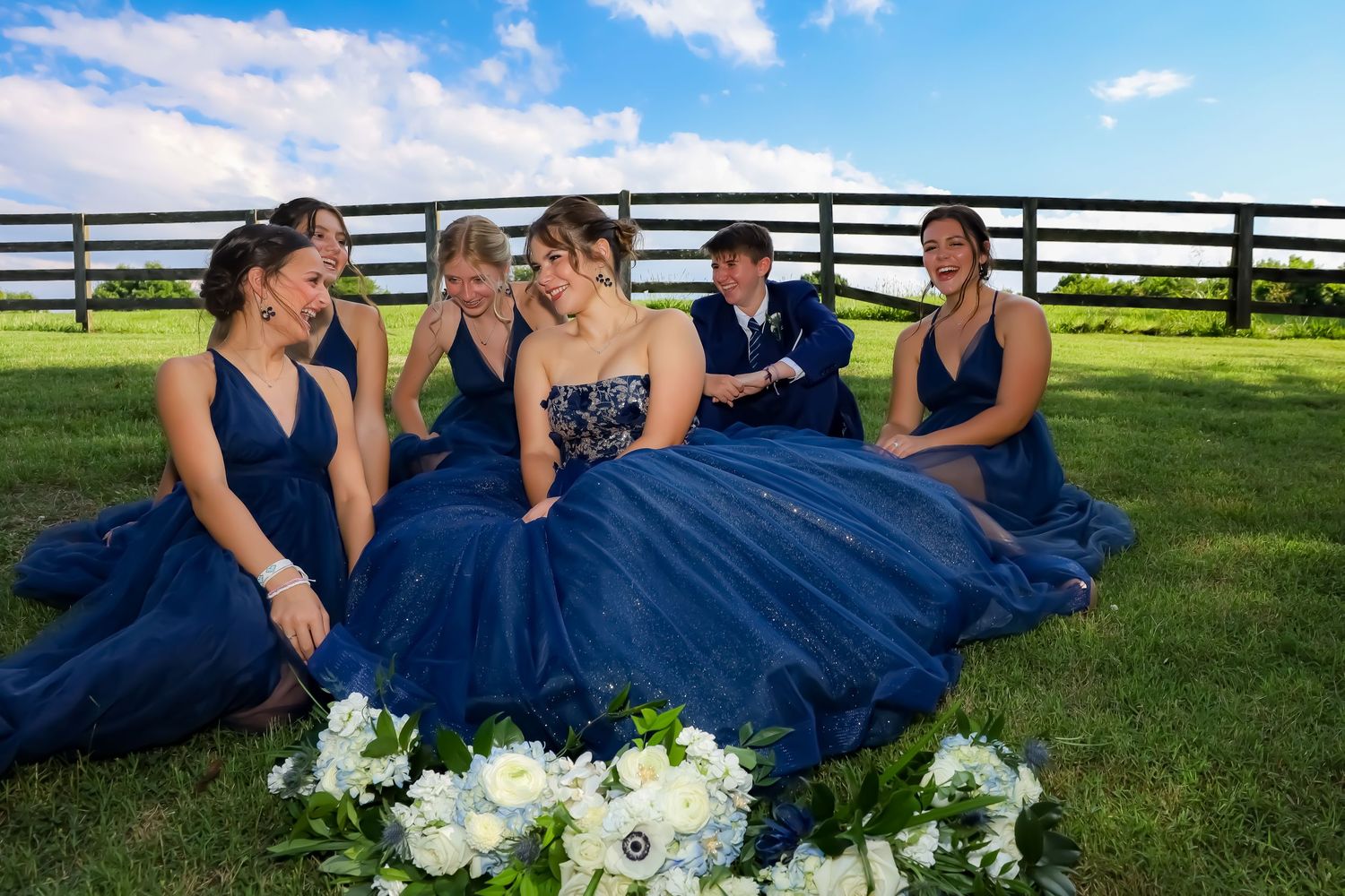 Bridesmaids in navy blue dresses sitting on grass with white floral bouquets.