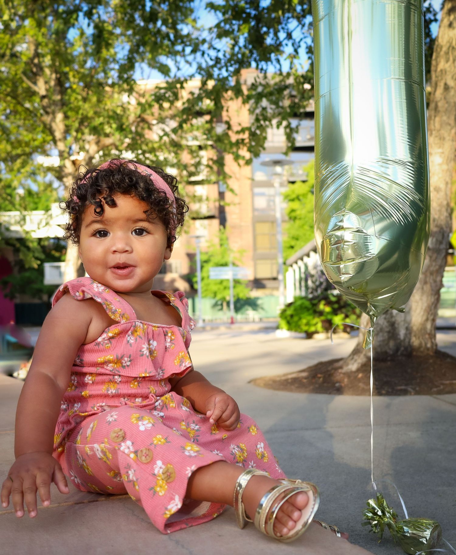 Baby in floral pink romper sitting on concrete ground near trees on sunny day.