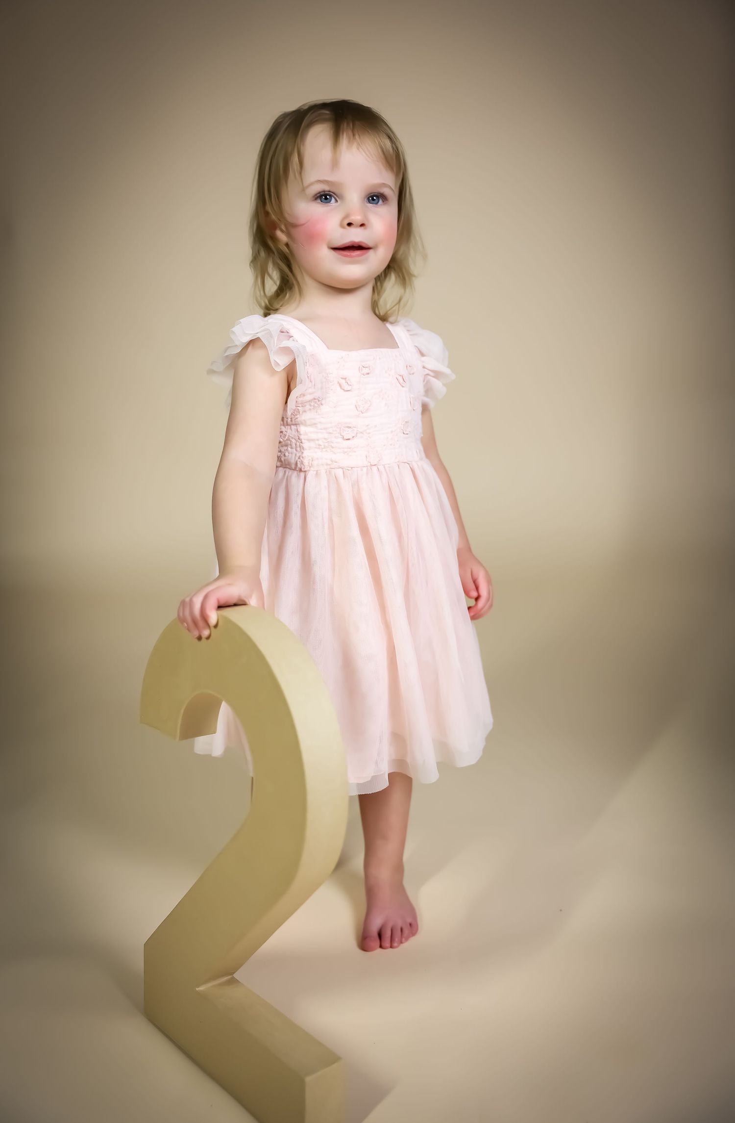 Toddler in pink dress standing beside gold number two for birthday photos.