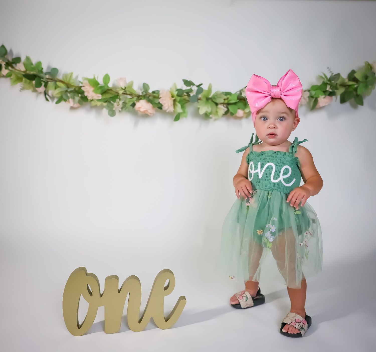 First birthday photo with mint green tulle dress and gold 'one' sign under floral garland.