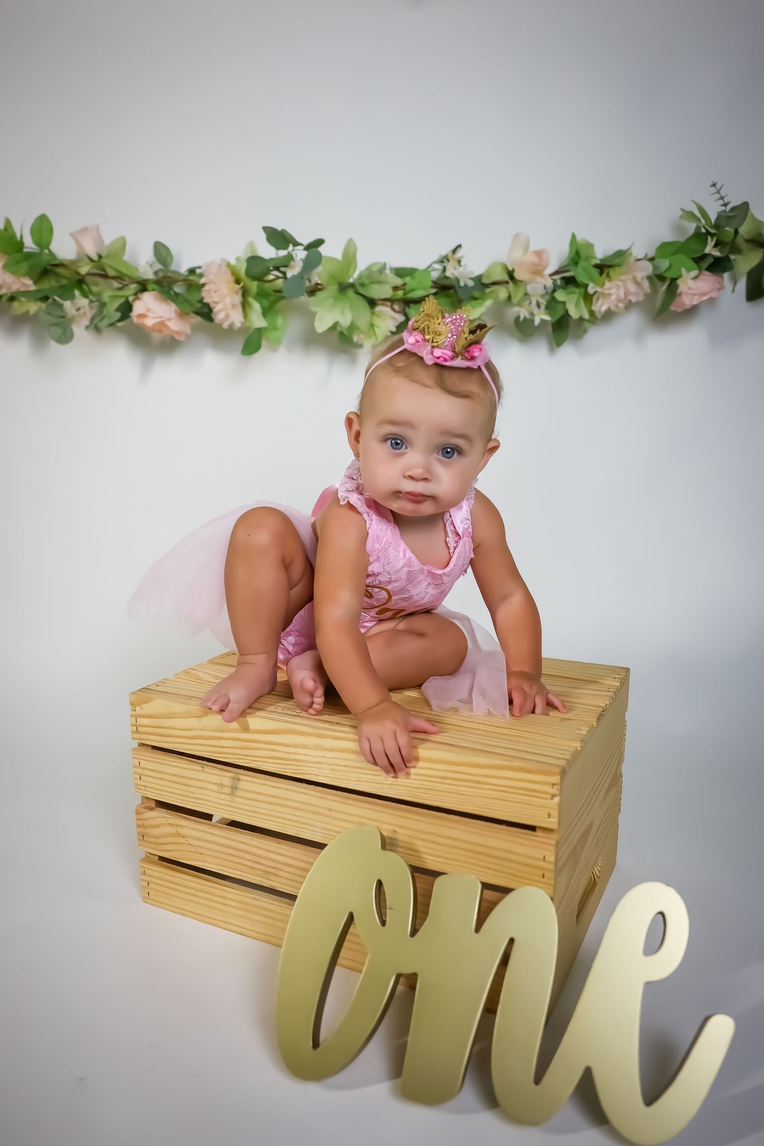 First birthday photo setup with floral garland and wooden crate against white background.