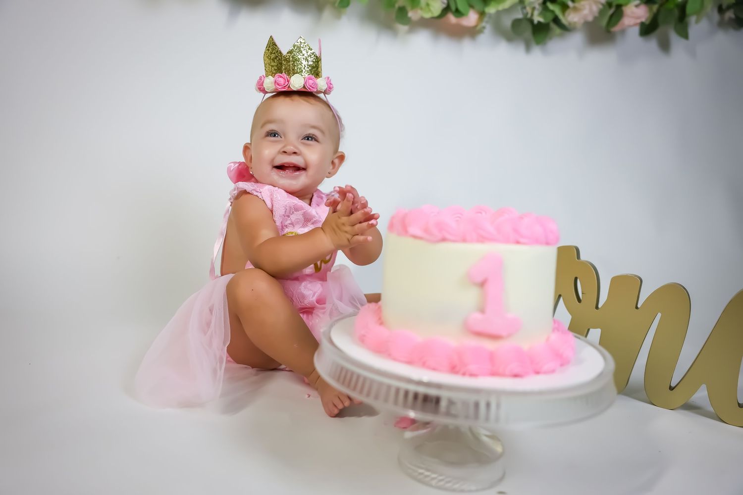 First birthday celebration with pink cake and gold decorations against white backdrop.