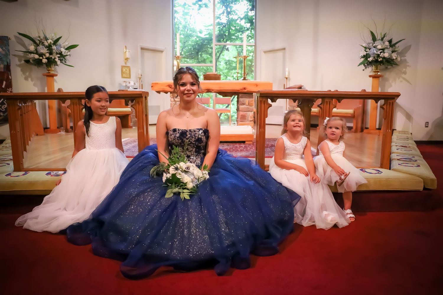 Wedding party in navy blue dresses seated together on church pews.